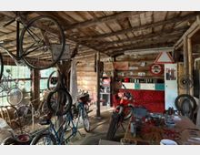 Holzwerkstatt mit rustikalem Interieur, Fahrrädern und Motorradteilen. An den Wänden hängen Verkehrsschilder, alte Kennzeichen und Werkzeuge; ein Arbeitstisch mit Ersatzteilen steht im Vordergrund.