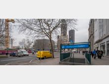Straßenszene am Kurfürstendamm in Berlin mit einem U-Bahn-Eingang, auf dem ein blaues Schild "Kurfürstendamm" zu sehen ist. Im Hintergrund befinden sich die Kaiser-Wilhelm-Gedächtniskirche, moderne Gebäude und ein Baukran, umgeben von Fußgängern und parkenden Autos.