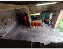 Ein rustikaler, offener Raum mit unverputzten Ziegelwänden, einer Bartheke mit Holzstühlen und einer Kühlschrank-Ecke. Im Hintergrund sind Dekorationen wie Poster, Schallplatten und Getränkeutensilien zu sehen, während durch die offene Tür Tageslicht einfällt und ein Blick ins Freie führt.