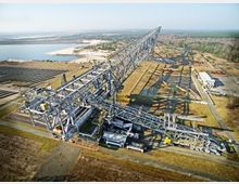 Luftaufnahme eines großen Tagebau-Baggers mit einer massiven Stahlkonstruktion, umgeben von einer weitläufigen Landschaft. Im Hintergrund sind ein See, Wälder und ein Industriegebiet mit Gebäuden sichtbar.