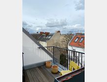 Dachterrasse mit Holzboden, schwarzem Geländer und drei leeren Blumentöpfen, im Hintergrund umliegende Gebäude mit roten und grauen Dächern sowie bewölkter Himmel.
