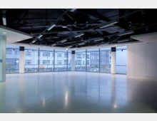 Großer, leerer Innenraum mit glänzendem Boden, einer deckenhohen Fensterfront, die den Blick auf ein gegenüberliegendes Gebäude mit mehreren Etagen ermöglicht, und einer schwarzen, sichtbaren Deckenkonstruktion mit Beleuchtungselementen.