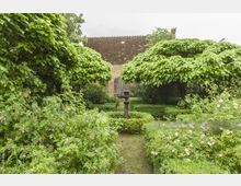 Ein gepflegter Garten mit symmetrischen Buchsbaumhecken, üppigem Grün und blühenden Sträuchern, im Hintergrund ein Backsteinhaus mit schrägem Dach und einer Holztür. In der Mitte des Gartens steht ein dekorativer Metallpfosten.
