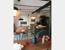 Ein moderner, gemütlicher Essbereich mit Holztischen und Stühlen, dekoriert mit Blumen in einer blauen Vase. Im Hintergrund befindet sich eine offene Küche mit einem Tresen, Geräten und einem Kühlschrank, sowie modernen Hängelampen und minimalistischem Dekor.