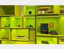 Ausstellungsraum mit leuchtend grünen Vitrinen, die retro-elektronische Geräte wie frühe Computer, Konsolen und Spiele zeigen, darunter ein Commodore C64, ein Game & Watch-Spiel und ein Space Wars-Spielautomat. Die Exponate sind in quadratischen, beleuchteten Fächern angeordnet.