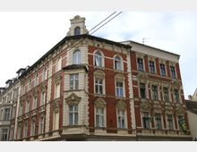 Eckgebäude im Stil des Historismus mit roter Klinkerfassade und hellen Stuckverzierungen, Rundbogenfenstern und Giebeldach. Im Erdgeschoss befinden sich Ladenschilder.