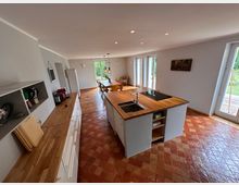 Moderne Küche mit zentraler Kochinsel, Holzarbeitsplatten und Waschbecken. Die Küche öffnet sich zu einem Essbereich mit einem Holztisch und Stühlen, große Fenster und Glastüren bieten Blick ins Grüne.