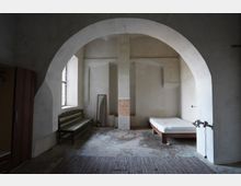 Spärlich eingerichteter, karger Innenraum mit großem Rundbogen, verputzten Wänden und rohem Betonboden, in der Mitte von einer massiven Stütze geteilt. Links stehen eine Holzbank und ein Fenster, rechts ein einfaches Bett; außerdem sind ein Spiegel und ein Schrank zu sehen.