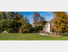 Parklandschaft mit einer großen Grünfläche im Vordergrund, umgeben von Bäumen in herbstlichen Farben. Im Hintergrund steht ein einfaches, zweistöckiges Gebäude mit grauer Fassade, teilweise von Graffiti verziert, daneben eine mit Efeu bewachsene alte Mauer.