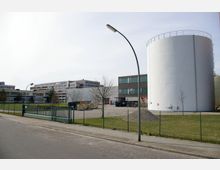 Großes Industriegelände mit mehreren modernen Gebäuden und einem großen weißen Zylinderbehälter rechts im Bild. Im Vordergrund sind ein Zaun, eine gepflasterte Straße und einige Bäume sichtbar.