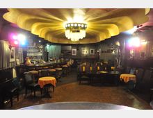 Innenraum einer Lounge oder Bar mit runder Deckenleuchte im Art-déco-Stil und wellenförmigen Verzierungen. Im Vordergrund stehen Tische mit gelben Tischdecken und schwarze Stühle, im Hintergrund befindet sich eine lange Bar mit Hockern, Regalen und dekorativen Elementen.