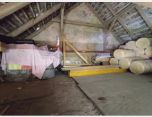 Dachboden mit offenem Holzdachstuhl, grobem Putz an den Wänden und gelagerten Baumaterialien, darunter große Rollen von Dämmstoff sowie Abdeckungen und Bauwerkzeuge. Der Boden besteht aus groben, teilweise schmutzigen Platten.