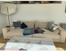 Wohnzimmer mit beigem Sofa, mehreren Kissen und einer karierten Decke; links steht eine weiße Stehlampe mit Holzstiel. Im Vordergrund liegt ein Couchtisch mit Weltkarten-Motiv, dahinter eine weiße Wand mit schmaler Ablage und Regalfach darüber.