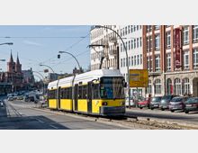 Eine gelb-schwarze Straßenbahn fährt durch eine städtische Straße in Berlin, links flankiert von der Oberbaumbrücke mit ihren markanten roten Türmen und rechts von mehrstöckigen Gebäuden mit Backsteinfassaden. Im Vordergrund sind Fahrbahnen, parkende Autos und Straßenschilder zu sehen.