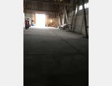 Innenansicht einer großen, offenen Lagerhalle mit unverputzten Ziegelwänden und sichtbaren Holzbalken. Am Ende der Halle ist ein großes, geöffnetes Tor mit einfallendem Tageslicht zu sehen, rechts und links stehen Baumaterialien und Geräte.