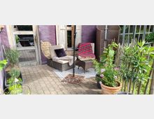 Kleiner gemütlicher Sitzbereich auf einer gepflasterten Terrasse mit lilafarbenem Haus im Hintergrund, ausgestattet mit zwei Rattan-Sesseln, einem Tisch, einem gemusterten Teppich und Dekokissen. Umgeben von Pflanzen in Töpfen und einem Holzzaun, ergänzt durch eine rustikale Holzfenstertür und eine teils freiliegende Backsteinmauer.