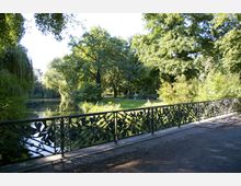 Ein parkartiger Bereich mit einem Teich, umgeben von hohen, dicht belaubten Bäumen. Im Vordergrund verläuft ein ornamentales, schmiedeeisernes Geländer mit floralen Mustern entlang eines gepflasterten Weges.