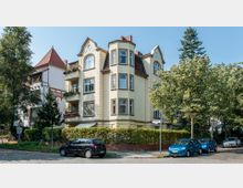 Eckgebäude im Stil eines Altbaus mit cremefarbener Fassade, Erkern und roten Dachschindeln, umgeben von Bäumen und einer niedrigen Hecke. Im Vordergrund geparkte Autos auf einer gepflasterten Straße.