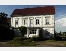 Zweistöckiges Wohnhaus mit weißer Fassade und roten Dachziegeln, bestehend aus symmetrischen Fenstern mit Rollläden sowie einem kleinen Anbau mit großen Fenstern. Vor dem Gebäude befindet sich ein Garten mit gepflegter Vegetation.