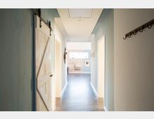 Langer, heller Flur in einem modernen Wohngebäude mit glatten Wänden in hellen Blau- und Weißtönen, einer Schiebetür aus hellen Holzplanken links, Hakenleiste rechts und einer Sicht auf ein Fenster und Möbel im Hintergrund.