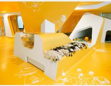 Innenraum eines bunten Kinderbereichs mit leuchtend gelben Wänden und Böden, verziert mit weißen Illustrationen von Tieren und Fantasiefiguren. Es gibt eine Kinderspielecke mit einer Ballgrube, ausgestopften Tieren und geschwungenen weißen Elementen, die moderne, spielerische Strukturen bilden.