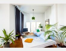 Helles Schlafzimmer mit zwei Einzelbetten, weißen Bettlaken und bunten Kissen, umgeben von großen Topfpflanzen. Im Hintergrund stehen ein kleiner Fernseher, ein Sessel sowie eine Fensterfront mit weißen Vorhängen und dunklen Gardinen.