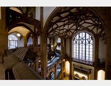 Innenansicht eines historischen Gebäudes mit gotischer Architektur, gekennzeichnet durch verzierte Spitzbögen, filigrane Gewölbe und große Buntglasfenster. Eine geschwungene Treppe und balustradengesäumte Gänge dominieren den Raum.