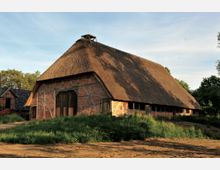 Historisches Fachwerkgebäude mit Backsteinwänden und einem großen Reetdach, umgeben von grüner Vegetation und einem sandigen Weg; dahinter sind weitere kleinere Gebäude und Bäume sichtbar.