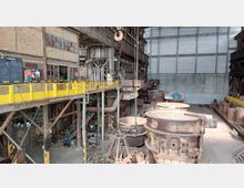 Industriehalle mit einer Stahlwerksanlage, einschließlich großer, rostiger Gusstiegel im Vordergrund und Laufstegen mit gelbem Geländer. Im Hintergrund sind Metallstrukturen, Rohrleitungen und eine Ziegelwand mit Fenstern und Tafeln erkennbar.