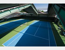 Zwei überdachte Tennisplätze mit blauem Spielfeld und grüner Umrandung in einer modernen Halle mit Stahlkonstruktionen; seitlich sind Fenster und mehrere Sitzbänke sichtbar.