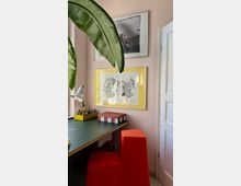 Ein kleiner Arbeitsplatz mit einer grünen Tischplatte, einer roten modernen Stuhlform und einer gelben Aufbewahrungsbox für Stifte. An der rosafarbenen Wand hängen zwei gerahmte Kunstwerke, eines davon mit gelbem Rahmen. Eine große Zimmerpflanze ragt in den Vordergrund.