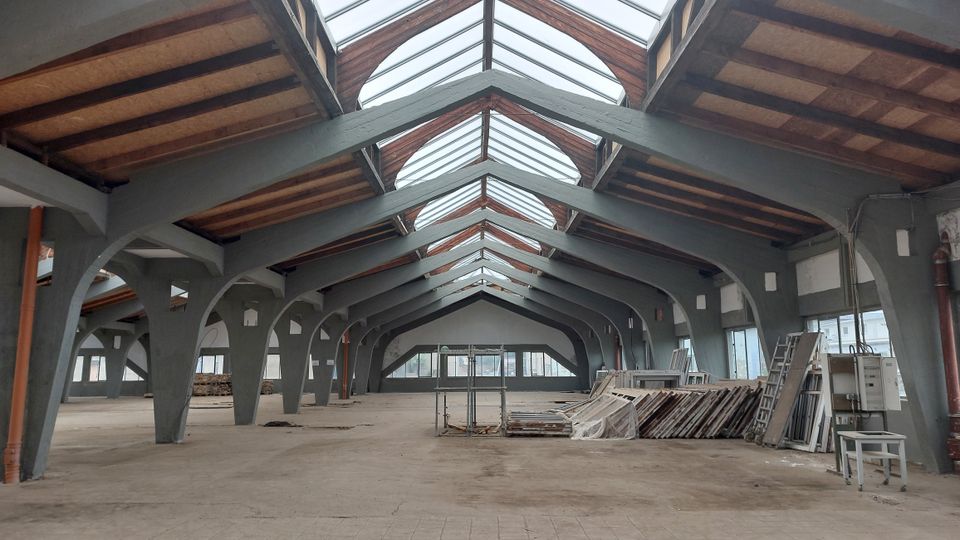 Geräumige, leerstehende Halle mit sichtbaren Gitterträgern aus Metall und Holzelementen, einem hohen Glasdach und großen Fenstern an den Seiten; Baumaterialien sind unordentlich in einem Bereich gestapelt.