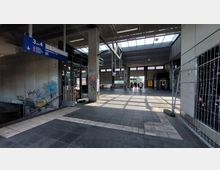 Offener Bereich eines Bahnhofs mit Betonwänden, Graffiti, hohen Fenstern und einem Glasdach, das Tageslicht einlässt. Im Hintergrund bewegen sich Menschen durch den Durchgang; auf der linken Seite sind Treppen und auf der rechten Seite Bauzäune sichtbar.