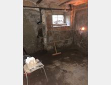 Ein leerer, einfacher Kellerraum mit unverputzten Wänden aus Ziegel und Beton sowie einem kleinen Fenster mit Gitter. Ein Besen lehnt an der Wand, Rohrleitungen verlaufen entlang der Decke, und ein Hocker mit Werkzeug und einer Farbeimer steht im Vordergrund auf einem abgenutzten Steinboden.