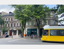 Straßenszene mit einem gelben Straßenbahnwagen im Vordergrund, der eine Haltestelle passiert. Im Hintergrund befinden sich historische Gebäude mit verzierten Fassaden, dahinter teilweise von Bäumen verdeckt, sowie ein Café mit Außensitzplätzen und geparkten Fahrrädern.
