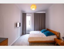 Schlafzimmer mit einem einfachen, modernen Design: ein Holzbett mit weißer Bettwäsche und blau-grünen Kissen steht rechts, daneben ein Nachttisch. Gegenüber hängt ein Flachbildfernseher an einer weißen Wand, und ein Fenster mit grauen Vorhängen sowie ein Heizkörper befinden sich am Ende des Raumes. Ein runder, gelblich leuchtender Deckenlampenschirm hängt in der Mitte.