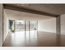 Großer, heller Raum mit Holzboden, einer Betondecke und weißen Wänden. Eine Wand besteht aus bodentiefen Fenstern, die den Blick auf eine Terrasse mit Pflanzen und ein Backsteingebäude ermöglichen.