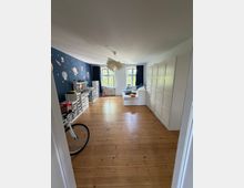 Heller Wohnraum mit breitem Holzdielenboden, zwei großen Fenstern mit dunklen Vorhängen und einer Deckenlampe. Links eine dunkelblaue Wand mit Weltraum-Tapete und Regalen, rechts eine Reihe weißer Kleiderschränke sowie ein weißes Tagesbett vor den Fenstern.
