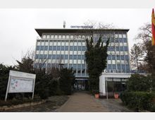 Modernes Gebäude mit der Aufschrift "Centre Français" auf dem Dach, umgeben von Bäumen und Sträuchern. Im Vordergrund führt ein gepflasterter Weg zum Eingangsbereich; daneben sind eine Informationstafel und eine deutsche Flagge zu sehen.