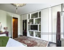 Schlafzimmer mit einer weißen Einbauschrankwand, die offene Regalfächer mit Büchern und Deko-Elementen sowie einen Fernseher enthält. Ein gemusterter Teppich bedeckt den dunklen Holzboden, und das Bett mit grüner Bettdecke befindet sich im Vordergrund.