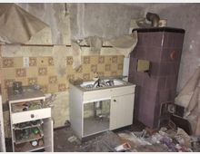 Eine alte, verlassene Küche mit beschädigten und abblätternden Tapeten, einem Spülbecken auf einem weißen Unterschrank, einem kleinen, offenen Regal mit Unordnung und einem violett gekachelten Ofen. Auf dem Boden liegt Müll und Schutt verstreut.