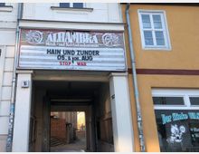 Ein Altbaugebäude mit einem Durchgang, darüber ein Schild mit der Aufschrift „ALHAMBRA Musik- und Kulturförderverein“ sowie Ankündigungen für Veranstaltungen und der Aussage „STOP WAR“. Rechts daneben ist eine gelbe Fassade mit einem Fenster und einem Ladenschild, auf dem „Der flinke Waschbär“ steht.