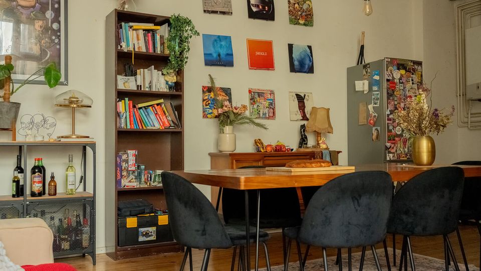 Eclectic dining room with a large wooden table and black upholstered chairs beneath hanging pendant bulbs. The space features a tall bookcase, a wall of framed art, a sideboard with decor, and a sticker-covered cabinet, with plants and a small bar shelf along the side.