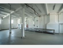 Industrieähnlicher Raum mit weiß gestrichenen Wänden und einer hohen Decke mit freiliegenden Rohren und Kabeln. Der Raum enthält mehrere Edelstahl-Arbeitstische, und an den Wänden sind eine Heizung, ein Boiler und Feuerlöscher sichtbar.