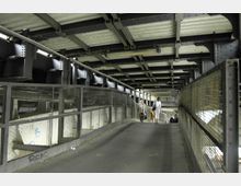 Überdachter Fußweg mit Metallgeländer und sichtbarer Stahlkonstruktion an der Decke. Zwei Personen gehen in entgegengesetzte Richtungen, und die Umgebung wirkt industriell mit beleuchteten Details.