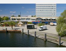 Hafenbereich mit Anlegestelle vor einem modernen Gebäude mit großen Fenstern und einer oberen Terrasse, auf der Sonnenschirme stehen. Im Vordergrund befinden sich Palmen, ein kleines Strohdachgebäude, ein Verkaufsanhänger und Lieferwagen, während Wasser und Poller im unteren Bildbereich sichtbar sind.