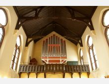 Innenraum einer Kirche mit Blick auf die Orgelempore. Sichtbar sind eine große Orgel in Rot und Weiß, ein hölzernes Geländer, dunkle Dachbalken und seitlich hohe Fenster mit Buntglas.