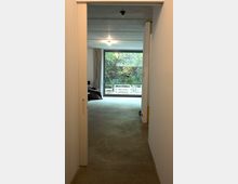 Blick durch eine offene Schiebetür in ein modernes Wohnzimmer mit Betonboden. Im Hintergrund ist ein großes Fenster mit Blick auf einen Garten mit Bäumen und Sträuchern sichtbar.