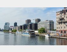 Städtische Uferlandschaft mit modernem Bürogebäude, Wohnhäusern und einem mit Graffiti bedeckten Industriebau rechts im Bild. Im Vordergrund liegt ein festgemachtes Boot, dahinter ein größeres Schiff mit der Aufschrift "Hostel & Lounge", am Wasser entlang sind Bäume und Spazierwege erkennbar.