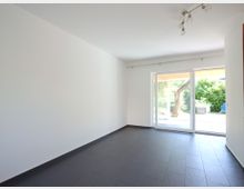 Leerer Raum mit weißen Wänden und dunklem Fliesenboden, ausgestattet mit einer Deckenlampe. Eine große Glasschiebetür führt zu einer Terrasse mit Blick auf einen Garten mit Bäumen und Sträuchern.